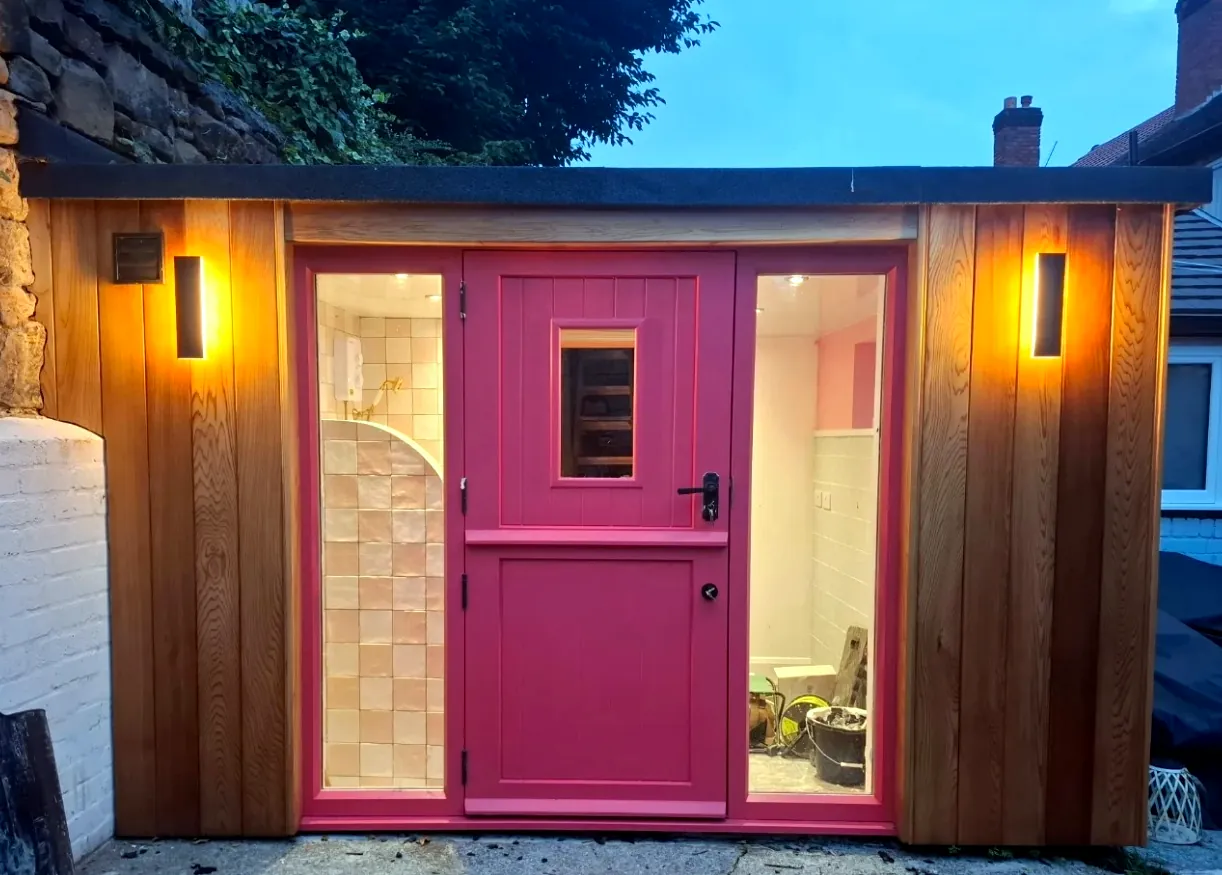 Cedar-clad garden room with a pink stable door, lit at dusk